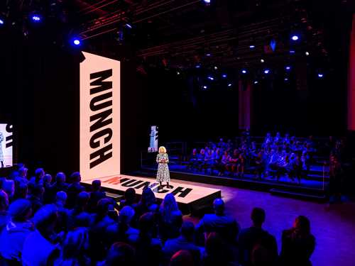 En stor sal med dempet blå belysning, i midten en kvinne som holder en presentasjon. 