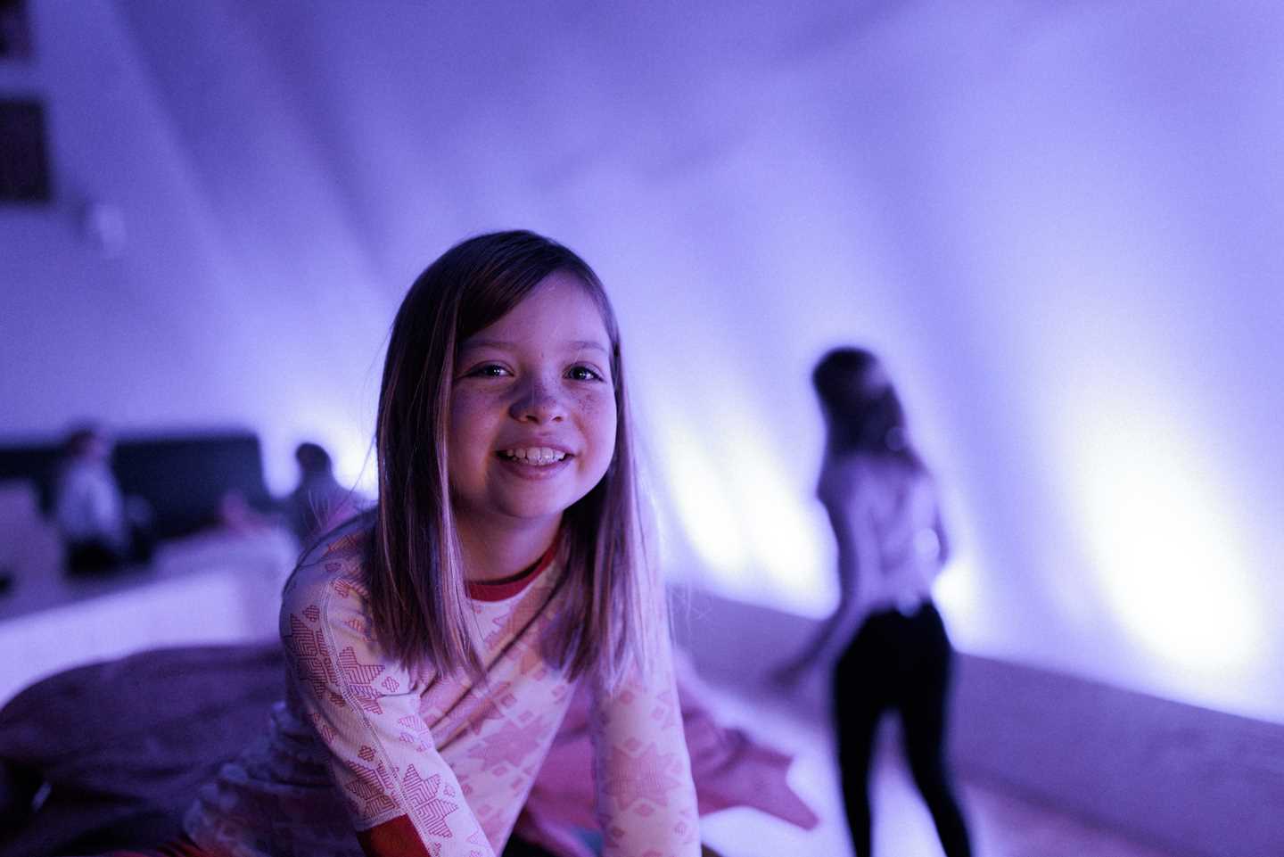 Barn leker i Kaoskammeret, i forgrunnen en jente som smiler og ser i kamera, omgitt av lilla lys og lekende barn i bakgrunnen ute av fokus
