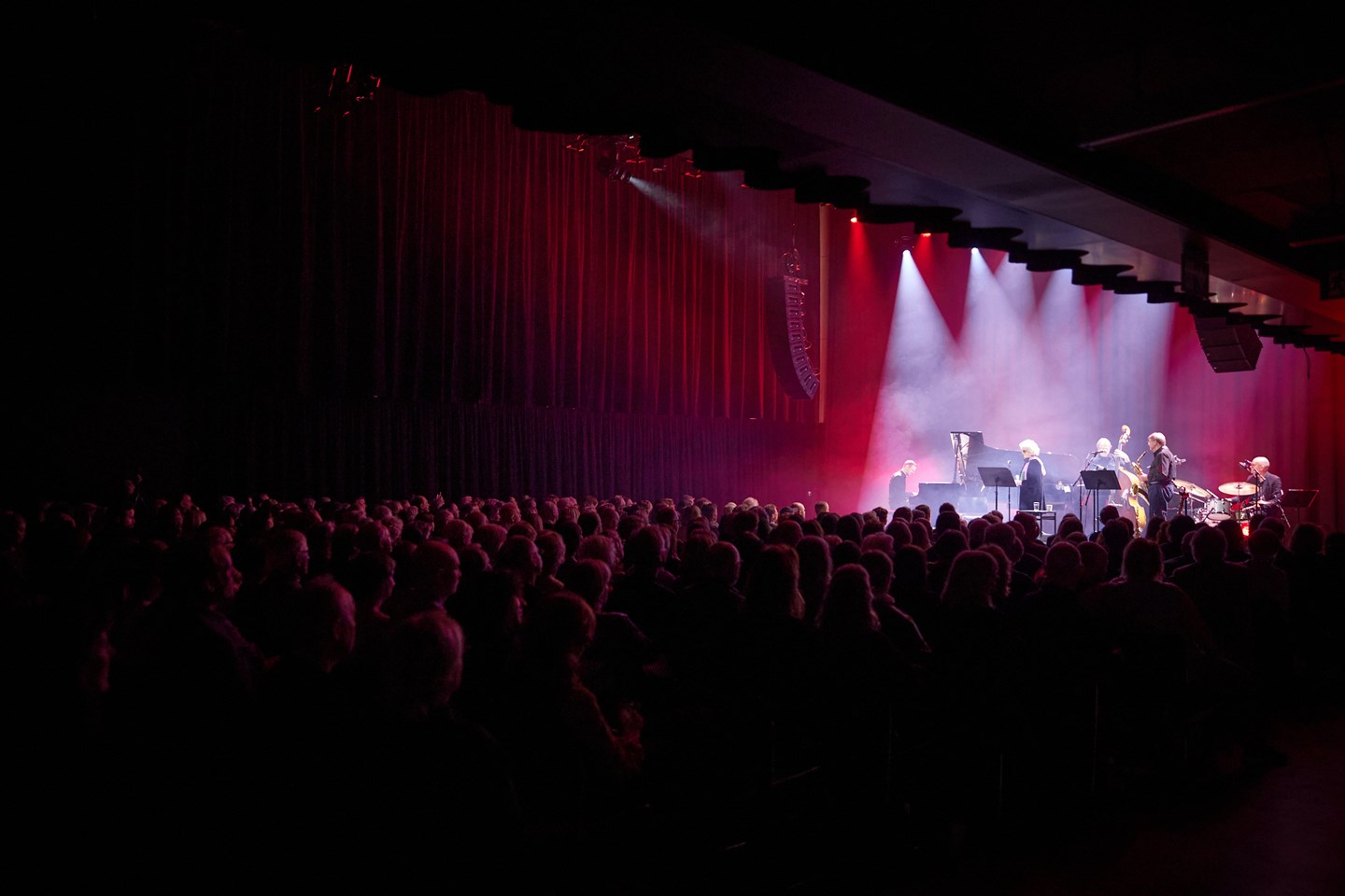 En fullsatt festsal omgitt av mørkt rosa lys, og spotlys mot scenen, hvor en gruppe jazzmusikere skimtes.